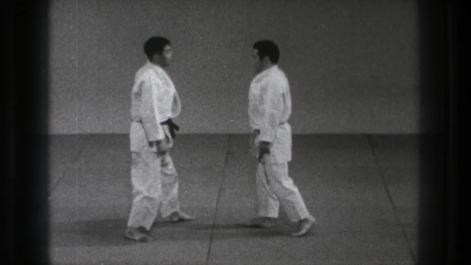 1971 Tokyo 8mm frame showing two adult karate practitioners in white gi approaching each other on a plain matted practice floor.