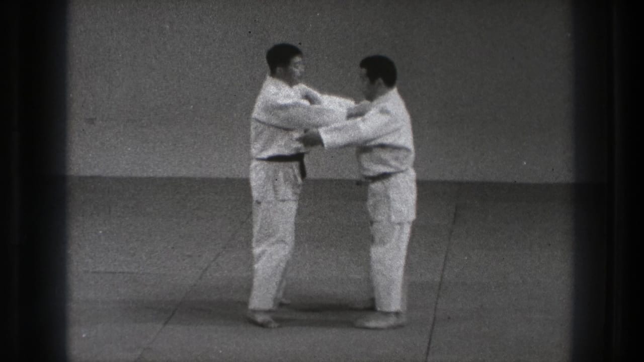 In 1971 Tokyo footage, two adult men in white gi and dark belts begin a controlled karate contact drill in a plain dojo.
