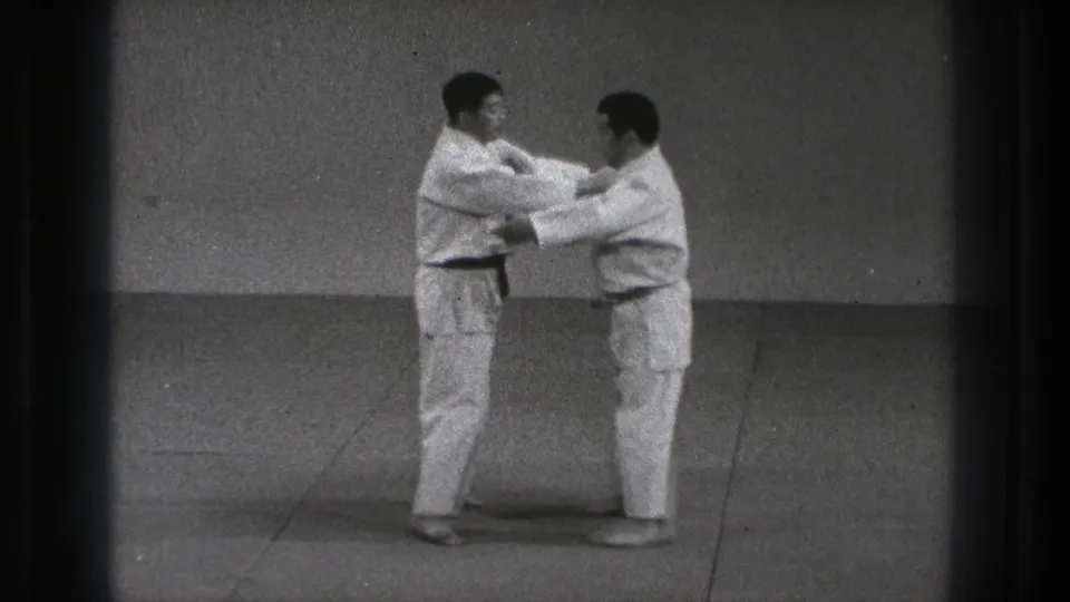 In 1971 Tokyo footage, two adult men in white gi and dark belts begin a controlled karate contact drill in a plain dojo.