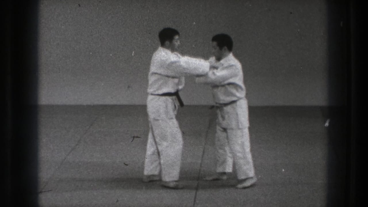 Two karate practitioners grip each other's sleeves or arms while standing on a mat.