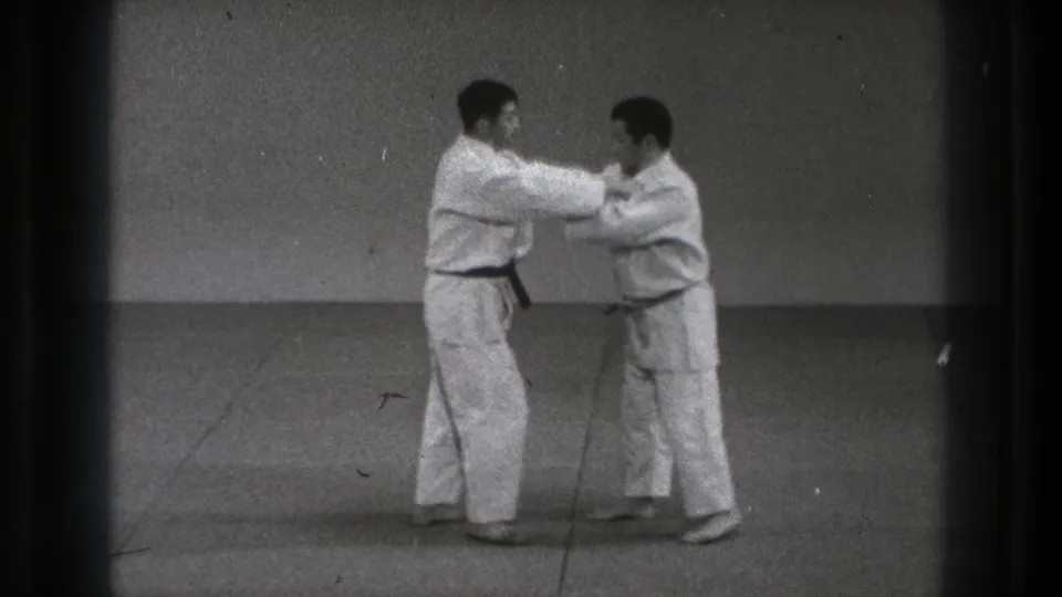 The two men maintain close contact during a measured karate or martial arts practice sequence in the simple Tokyo dojo setting.