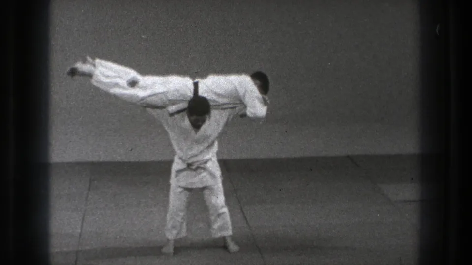 Tokyo 1971 8mm frame showing two adult men in white gi during a martial arts throw, with one practitioner lifted horizontally over the other.