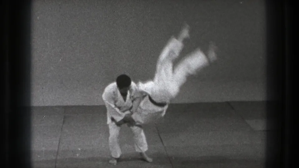 Tokyo 1971 8mm frame showing the throw at its most dynamic point, with one man in a gi rotating through the air above the mat.
