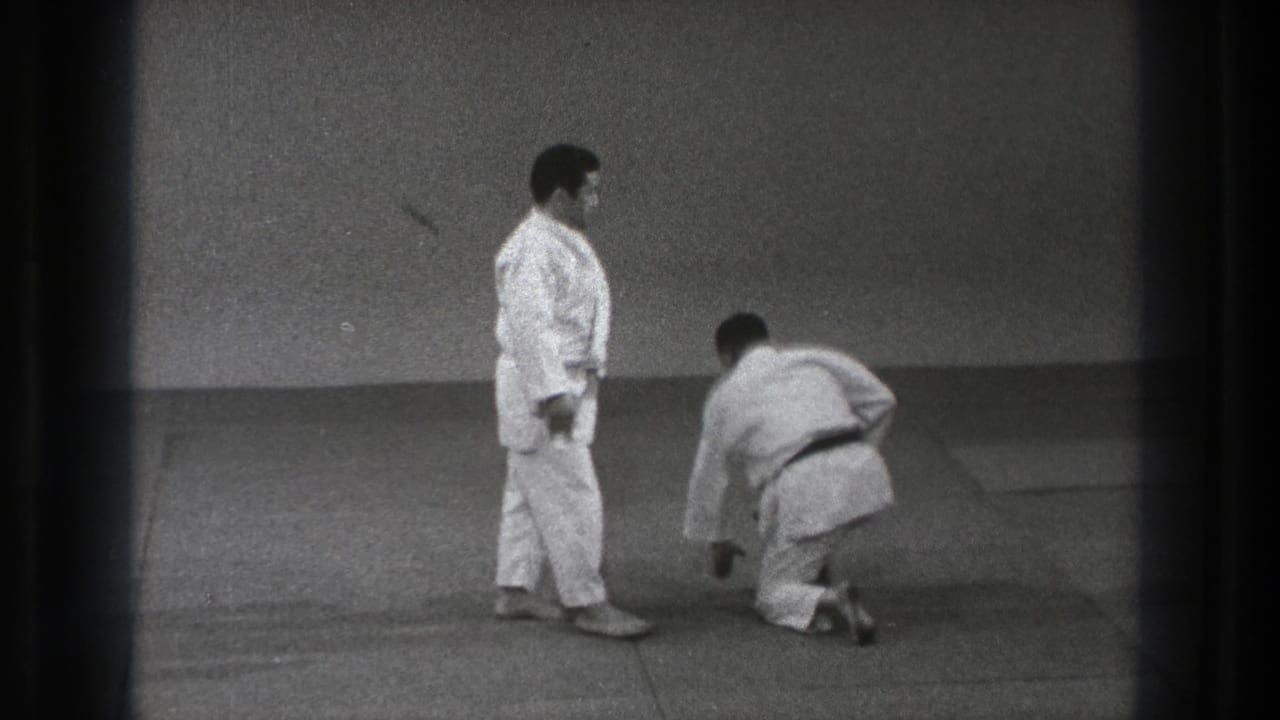 Two martial artists in gi on a mat, one kneeling while the other stands nearby.