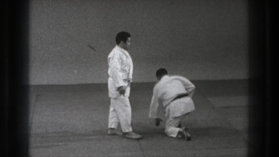 1971 Tokyo judo practice frame showing one gi-clad adult kneeling on the mat as another stands in a plain dojo setting.