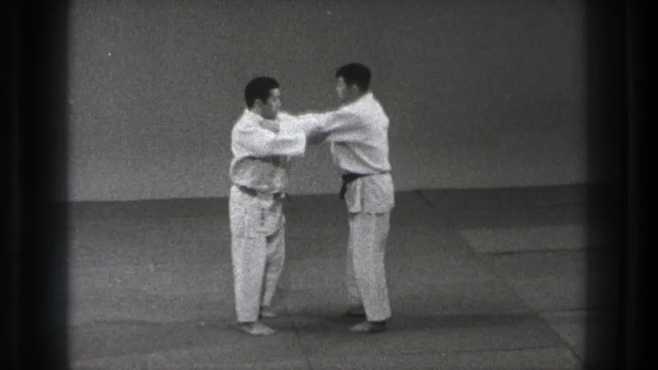 In supported 1971 Tokyo footage, two adult men in white judo uniforms stand in a grip position on a mat.