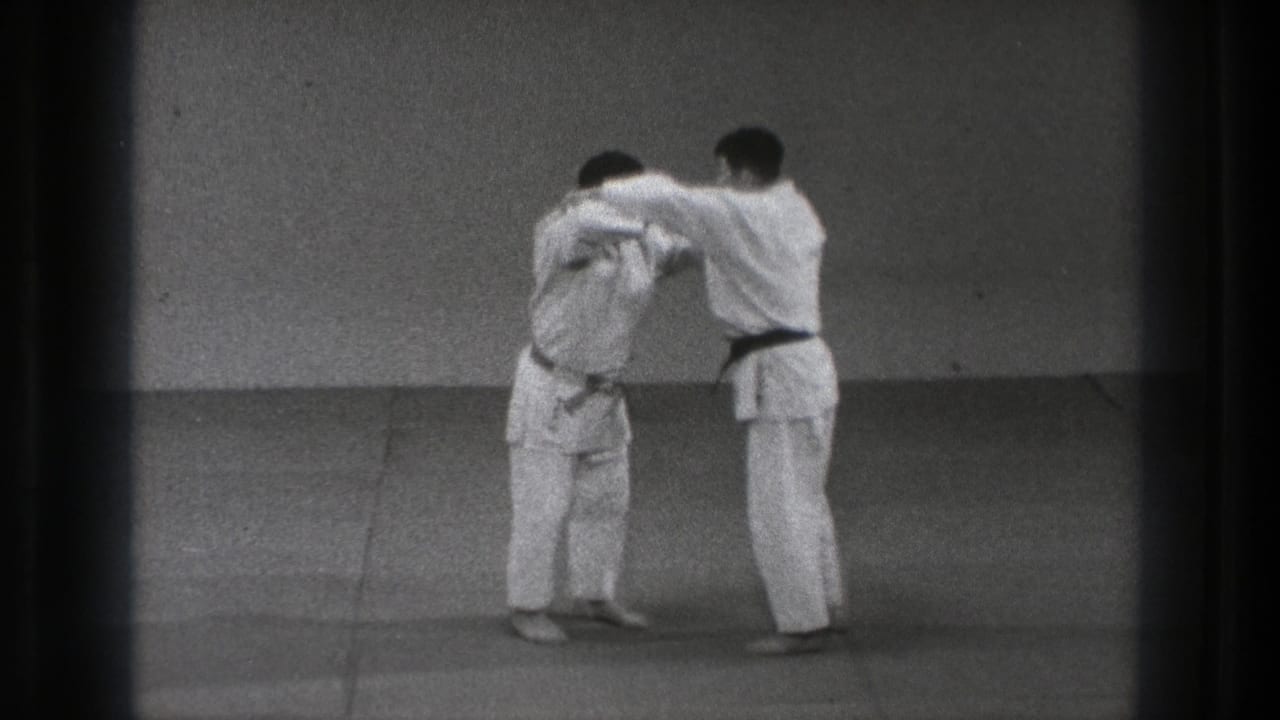 Two men in white uniforms continue a controlled judo grip sequence in the 1971 Tokyo training-room footage.