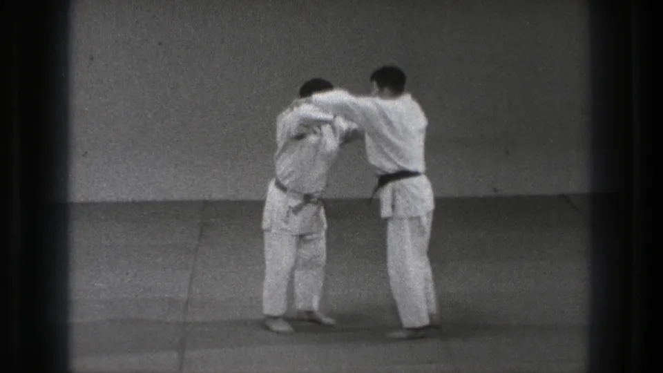 Two men in white uniforms continue a controlled judo grip sequence in the 1971 Tokyo training-room footage.