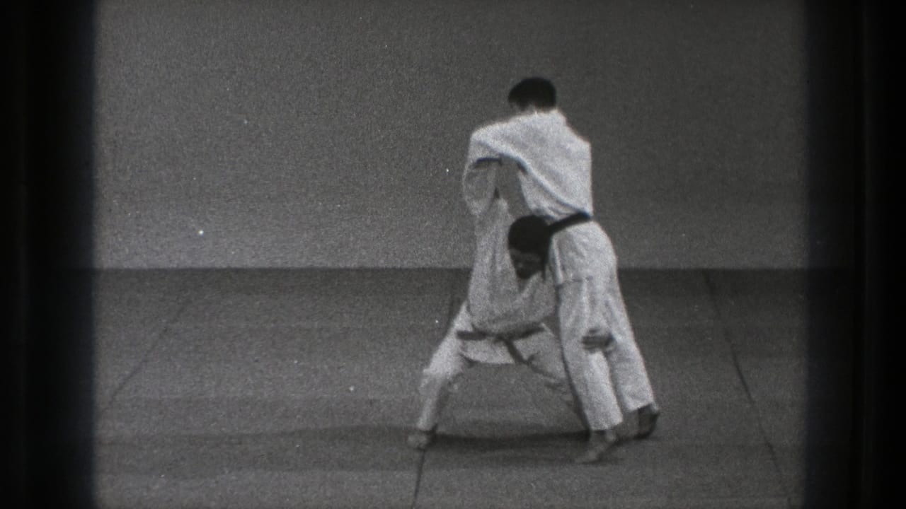 A black-belt practitioner positions his partner for a judo throw on the mat in the 1971 Tokyo clip.