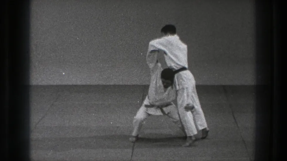 A black-belt practitioner positions his partner for a judo throw on the mat in the 1971 Tokyo clip.