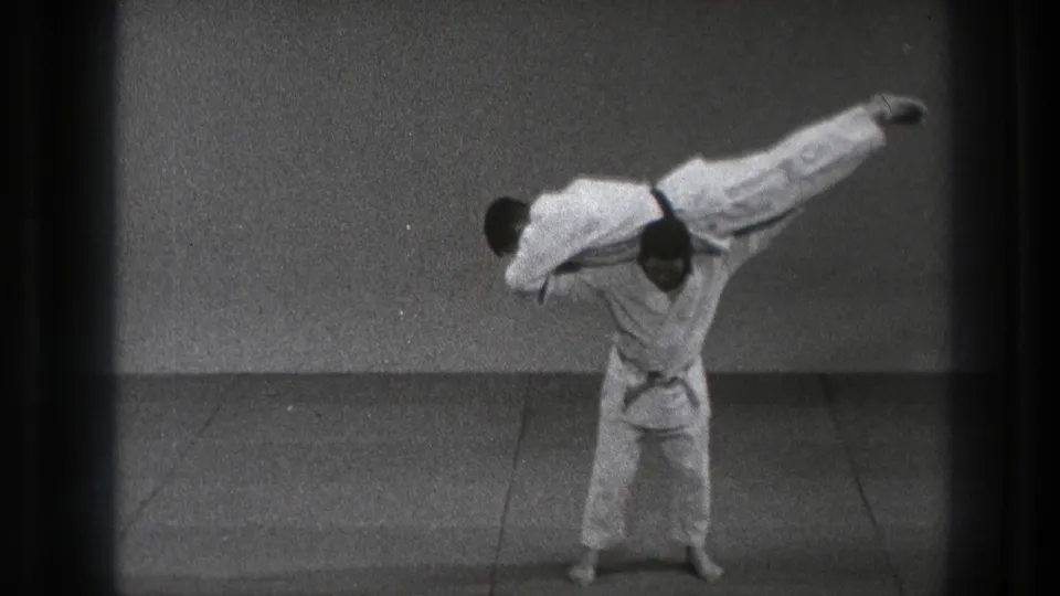 One man lifts another horizontally across his shoulders during a controlled judo throw demonstration in supported 1971 Tokyo footage.