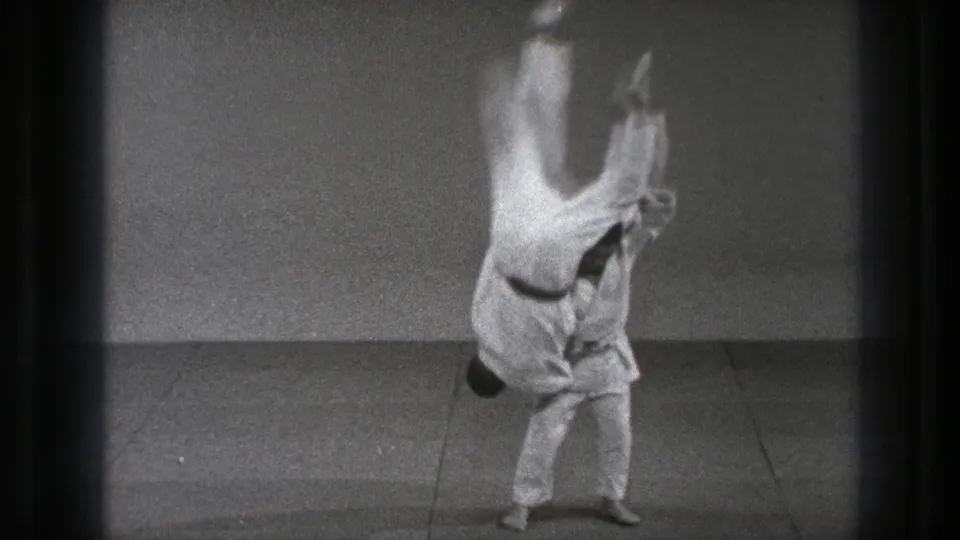 In 1971 Tokyo footage, one man in a white gi lifts another during a martial arts throw on a plain indoor mat.