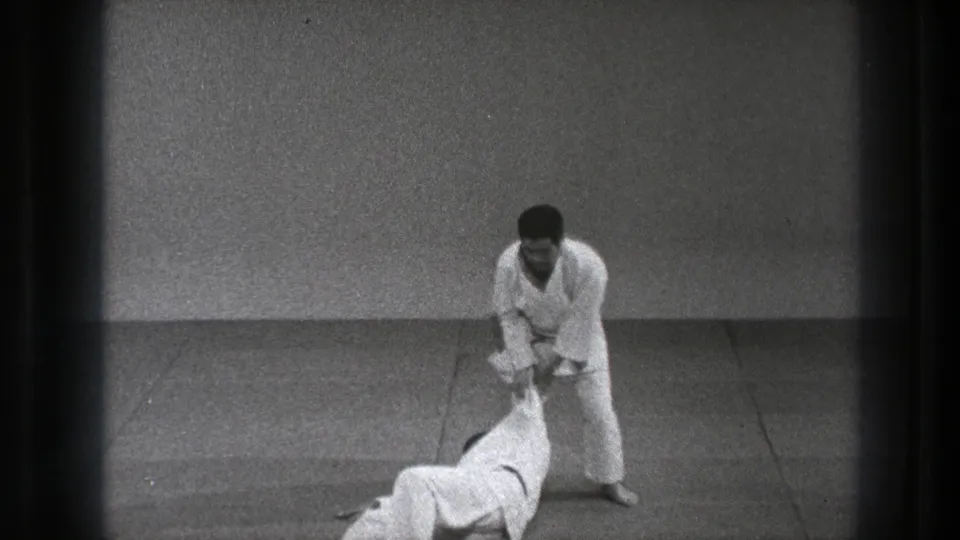 In the Tokyo martial arts sequence, one practitioner stands while the other lies on the mat after the throw.