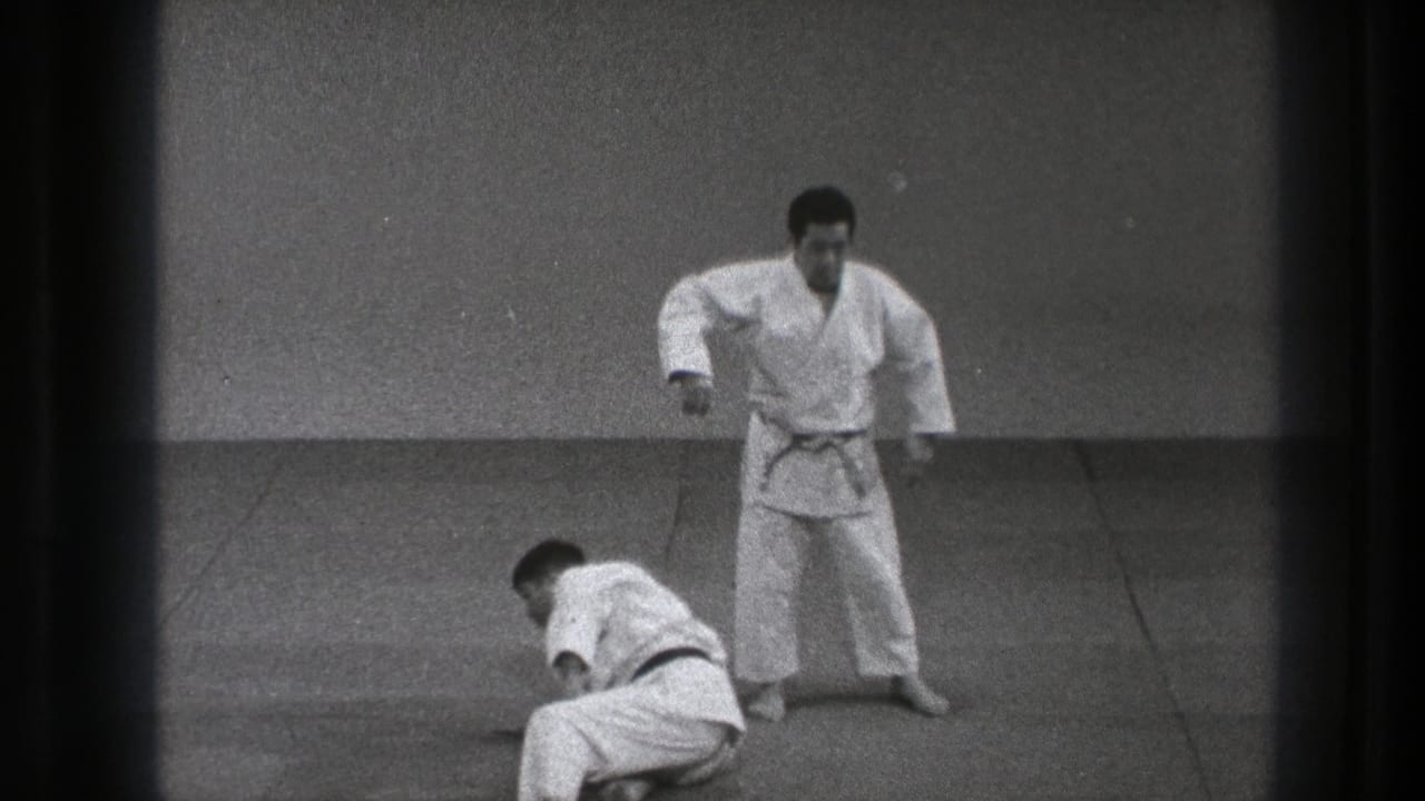 One adult practitioner in a white gi stands as his partner begins to recover from the mat in the 1971 Tokyo clip.