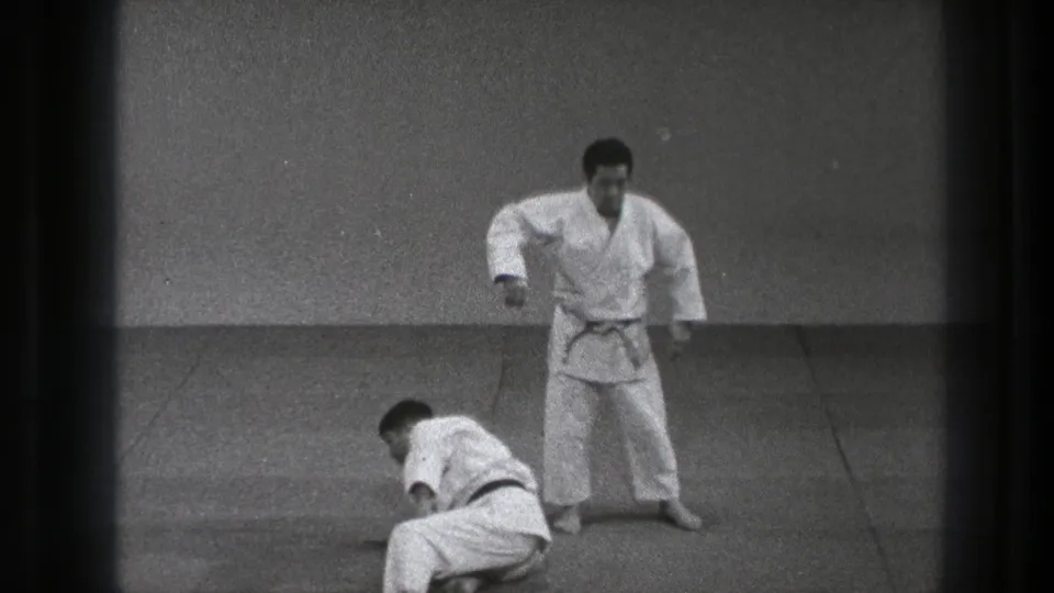 One adult practitioner in a white gi stands as his partner begins to recover from the mat in the 1971 Tokyo clip.