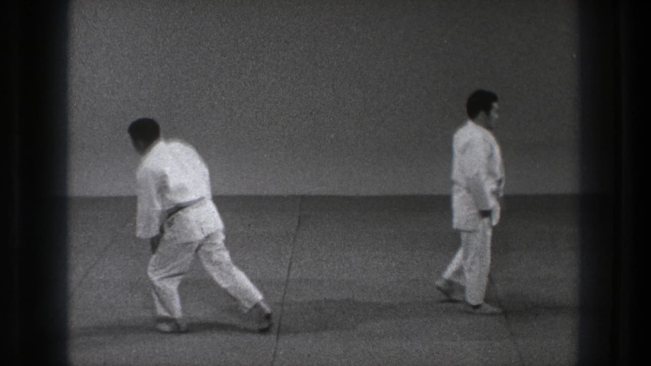 Two adult men in white gi uniforms and black belts move across a plain indoor floor in 1971 Tokyo footage.