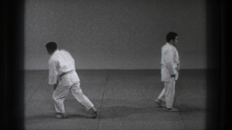 Two adult men in white gi uniforms and black belts move across a plain indoor floor in 1971 Tokyo footage.