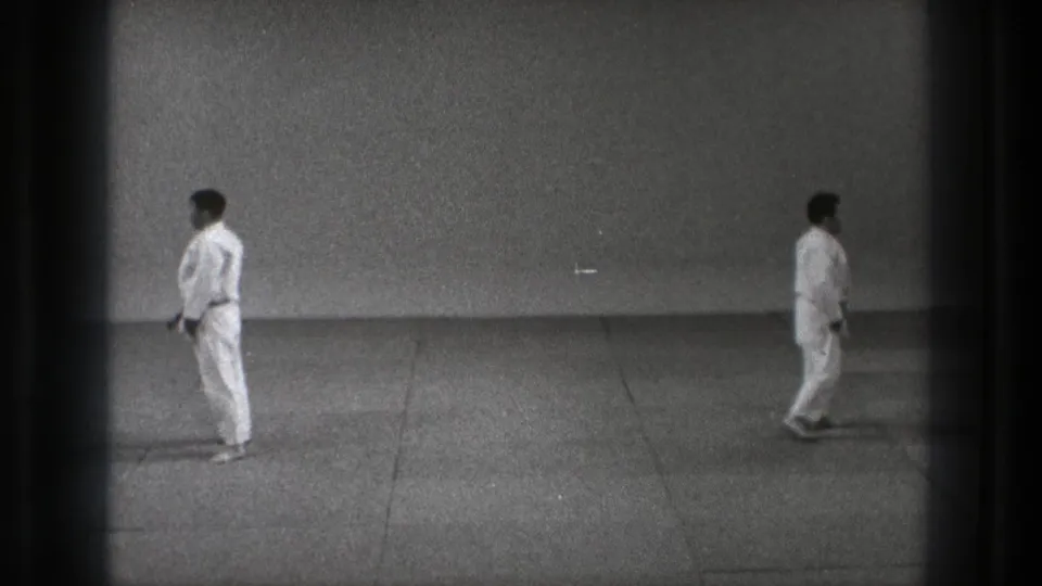 Two men in white gi uniforms and dark belts pause across the indoor floor in the 1971 Tokyo martial arts footage.