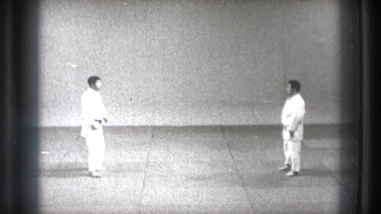 1971 Tokyo martial arts frame showing two adult men in white gi facing each other on grey mat flooring in a sparse indoor training space.