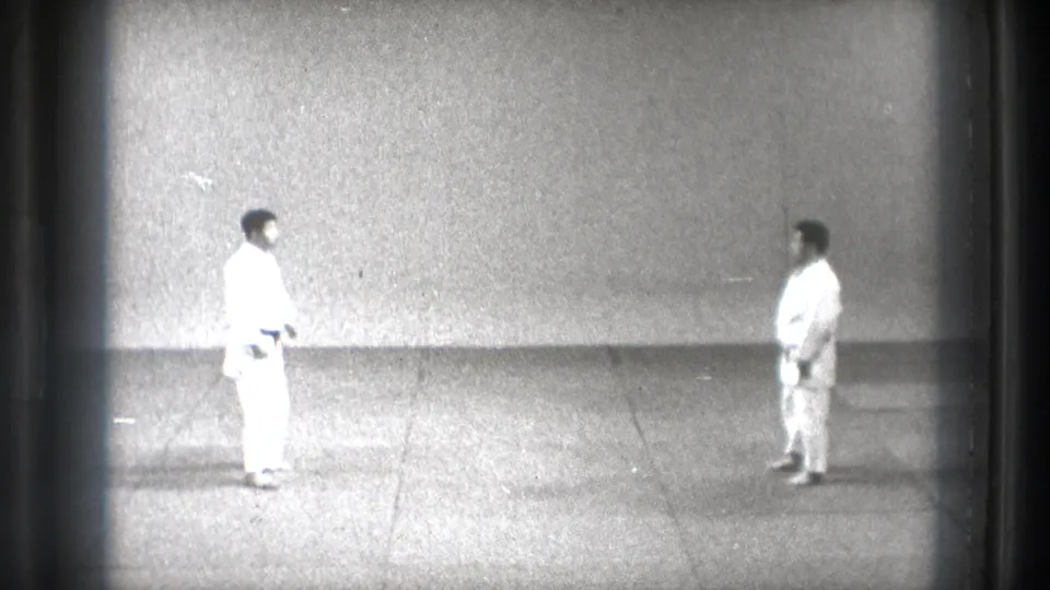 1971 Tokyo martial arts frame showing two adult men in white gi facing each other on grey mat flooring in a sparse indoor training space.