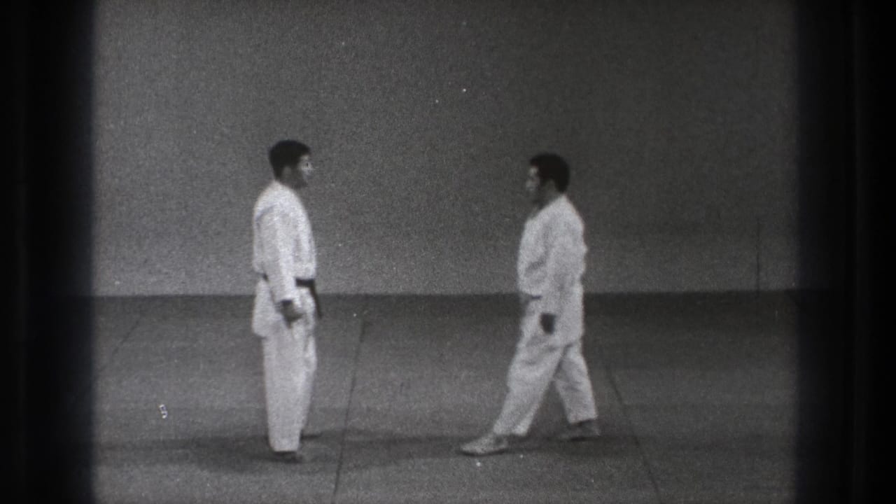 Two adult men in white judogi face each other in a Tokyo dojo setting, with one stepping closer across the mat.
