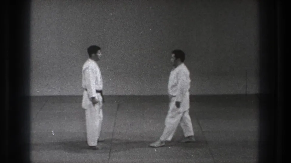 Two adult men in white judogi face each other in a Tokyo dojo setting, with one stepping closer across the mat.