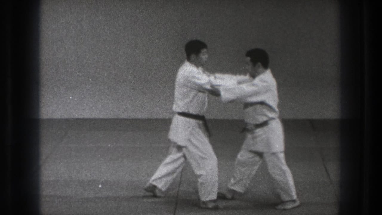Two men in karate uniforms face each other with hands engaged in a plain training room.