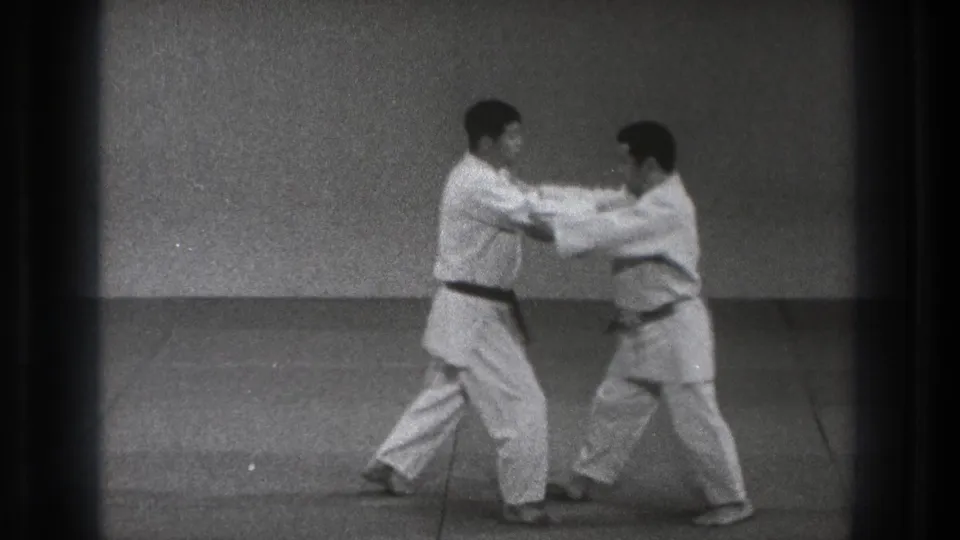 In supported 1971 Tokyo footage, two adult men in white karate gi practice a standing hand technique in a sparse indoor dojo setting.
