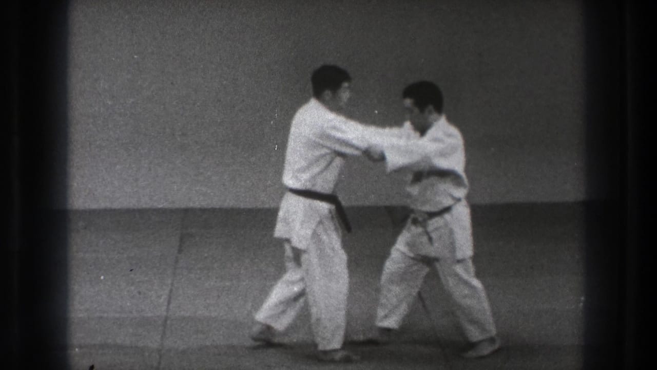 Two men in white gi maintain arm contact during a karate drill, with one practitioner in a black belt stepping forward.