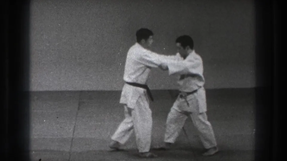 Two men in white gi maintain arm contact during a karate drill, with one practitioner in a black belt stepping forward.