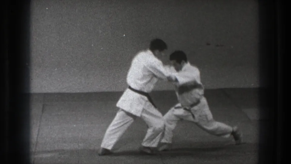 In the 1971 Tokyo martial arts clip, one man drops into a low stance as his partner controls the upper body during the technique.