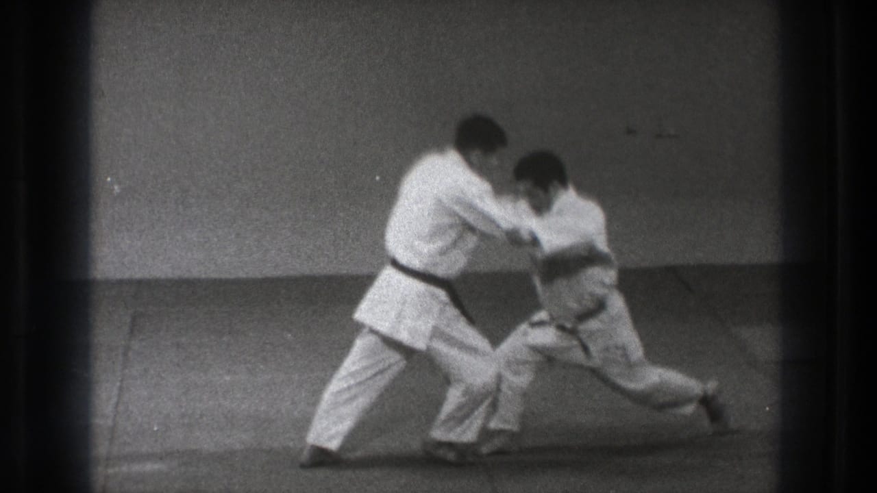 The partnered karate demonstration reaches a low, extended position, with both men in white gi framed by visible 8mm film edges.