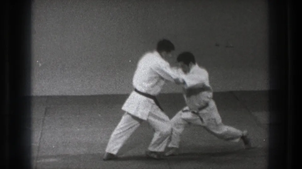 The partnered karate demonstration reaches a low, extended position, with both men in white gi framed by visible 8mm film edges.