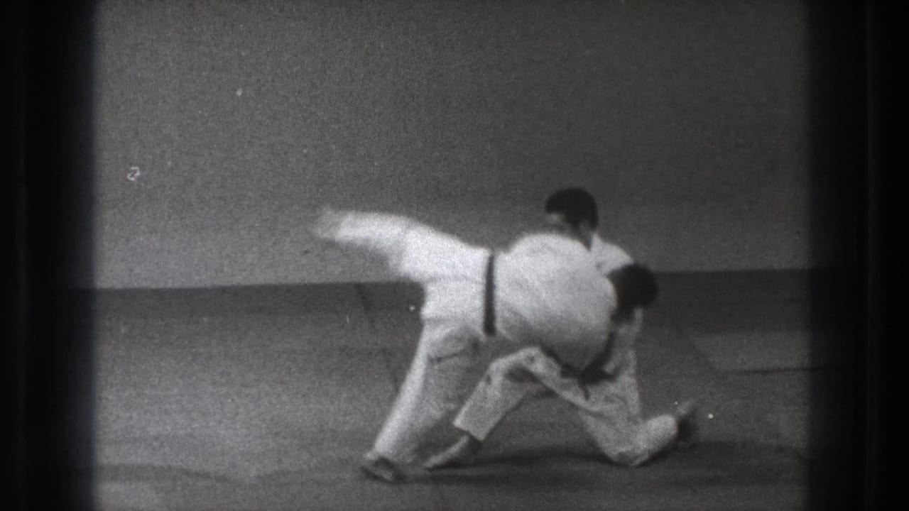 Two judoka in white gi begin a throw on a mat in a plain indoor room.