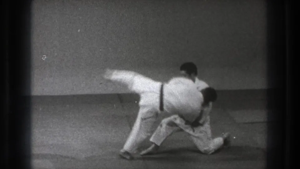 In 1971 Tokyo footage, two adult judoka in white gi and dark belts grapple low on a mat as one begins a throwing motion.