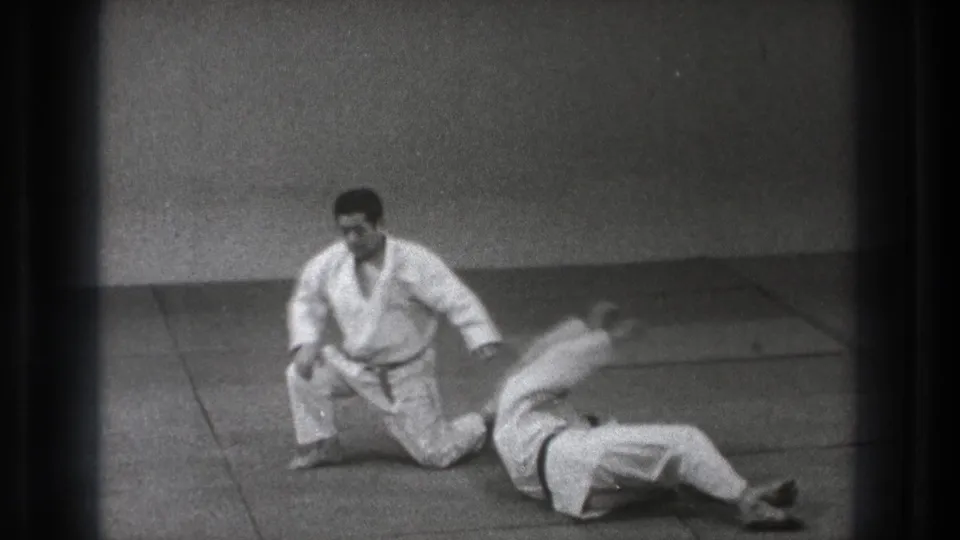 After the throw, one judoka kneels upright while his partner lies on the mat in a simple indoor Tokyo practice space.