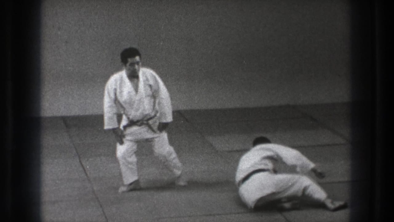 In 1971 Tokyo footage, two adult martial artists in white uniforms are on mats after a throw, with one standing and one seated on the floor.