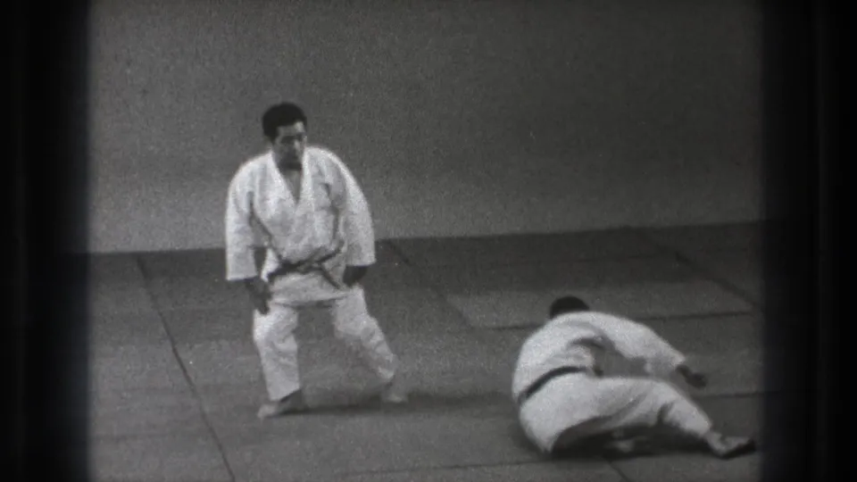 In 1971 Tokyo footage, two adult martial artists in white uniforms are on mats after a throw, with one standing and one seated on the floor.
