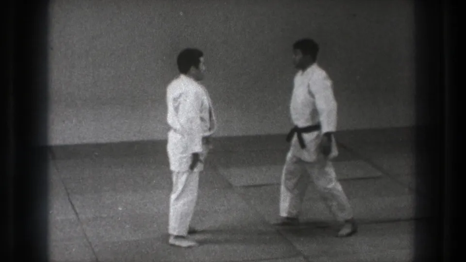 Two adult martial artists in white uniforms face each other on padded mats during a 1971 Tokyo practice sequence.