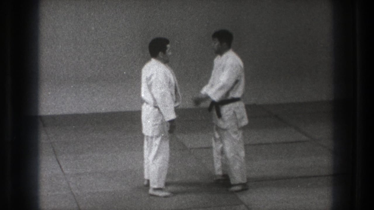 Two men in white martial arts uniforms stand facing each other on mats in a plain room.