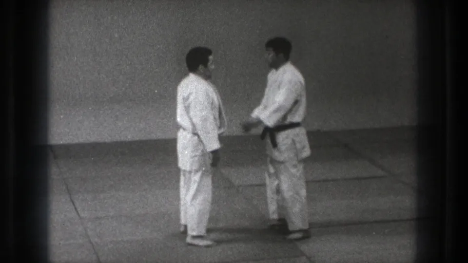 In 1971 Tokyo footage, two adult men in white gi face each other on a mat before beginning a martial arts drill.