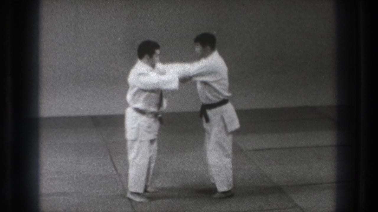 Two men in gi engage at arm's length during a 1971 Tokyo martial arts training exchange on indoor mats.