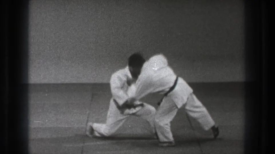 The exchange ends with one practitioner lunging low while the other bends forward in a takedown motion on the dojo mat.