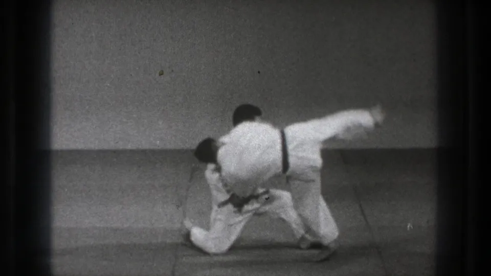 In 1971 Tokyo footage, one martial artist grips another while kneeling on a padded mat during a throw demonstration.