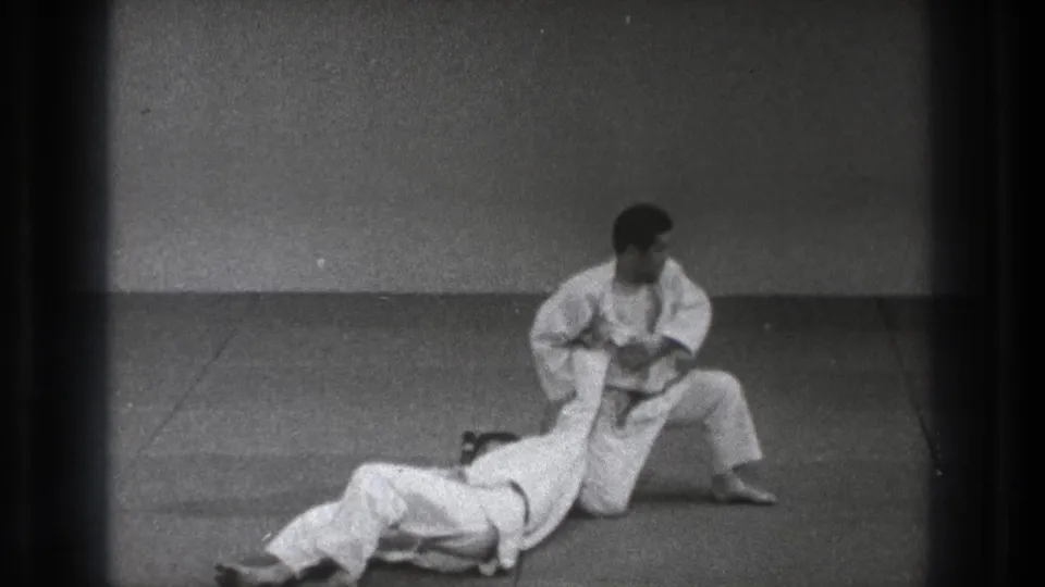 After the throw, the training partner lies on the mat while the kneeling martial artist maintains a controlled grip.