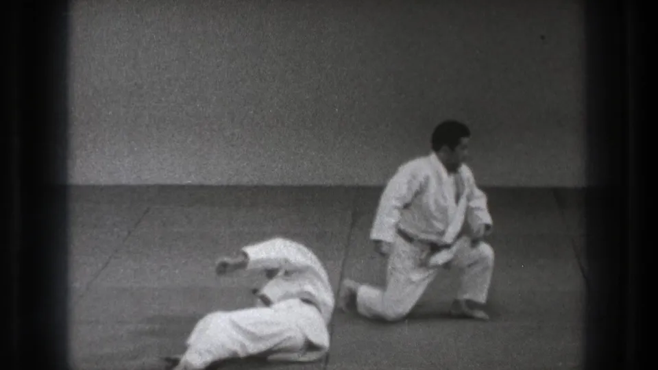 The demonstration ends with one martial artist kneeling beside his training partner, who remains on the mat in the indoor practice space.
