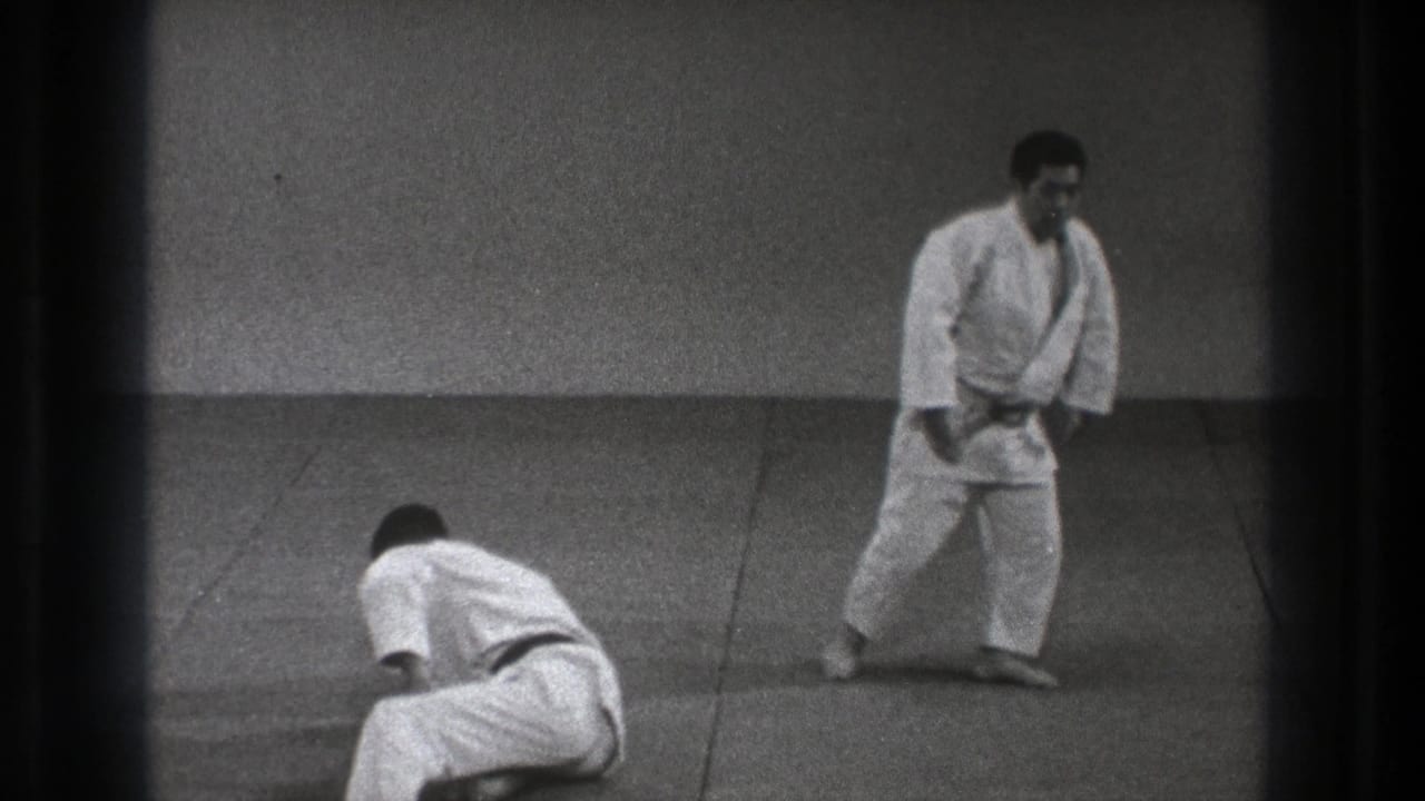 In 1971 Tokyo footage, two adult judo practitioners in white uniforms hold a demonstration position on a plain dojo mat.