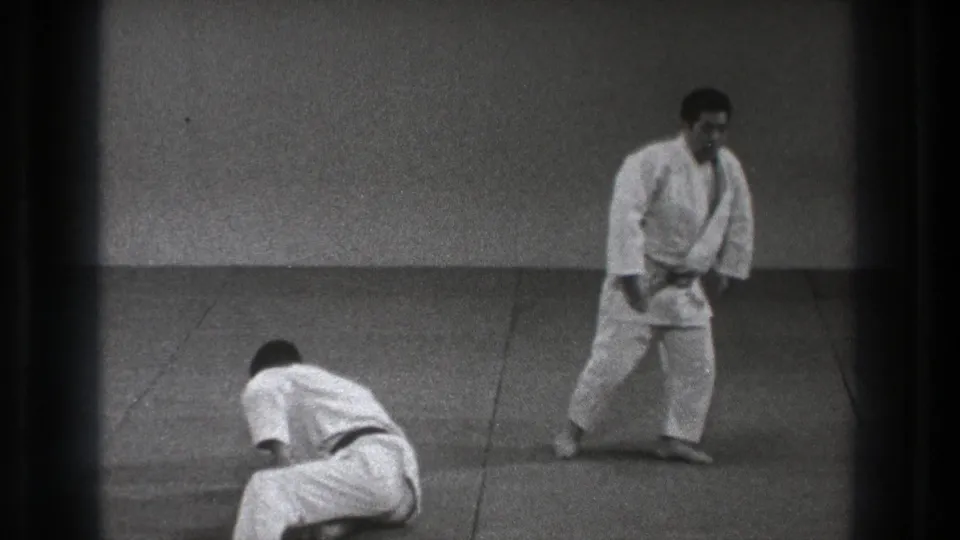 In 1971 Tokyo footage, two adult judo practitioners in white uniforms hold a demonstration position on a plain dojo mat.