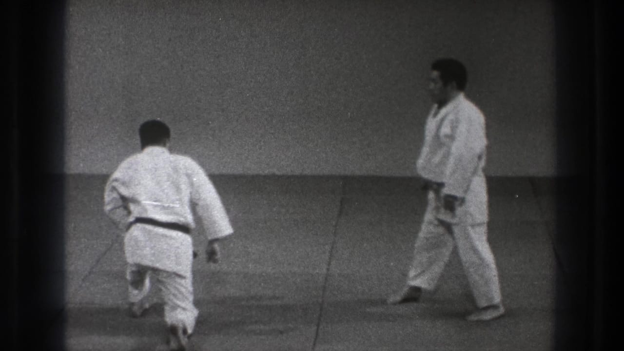 Two judo practitioners face each other on a mat, one moving forward from a kneeling position.