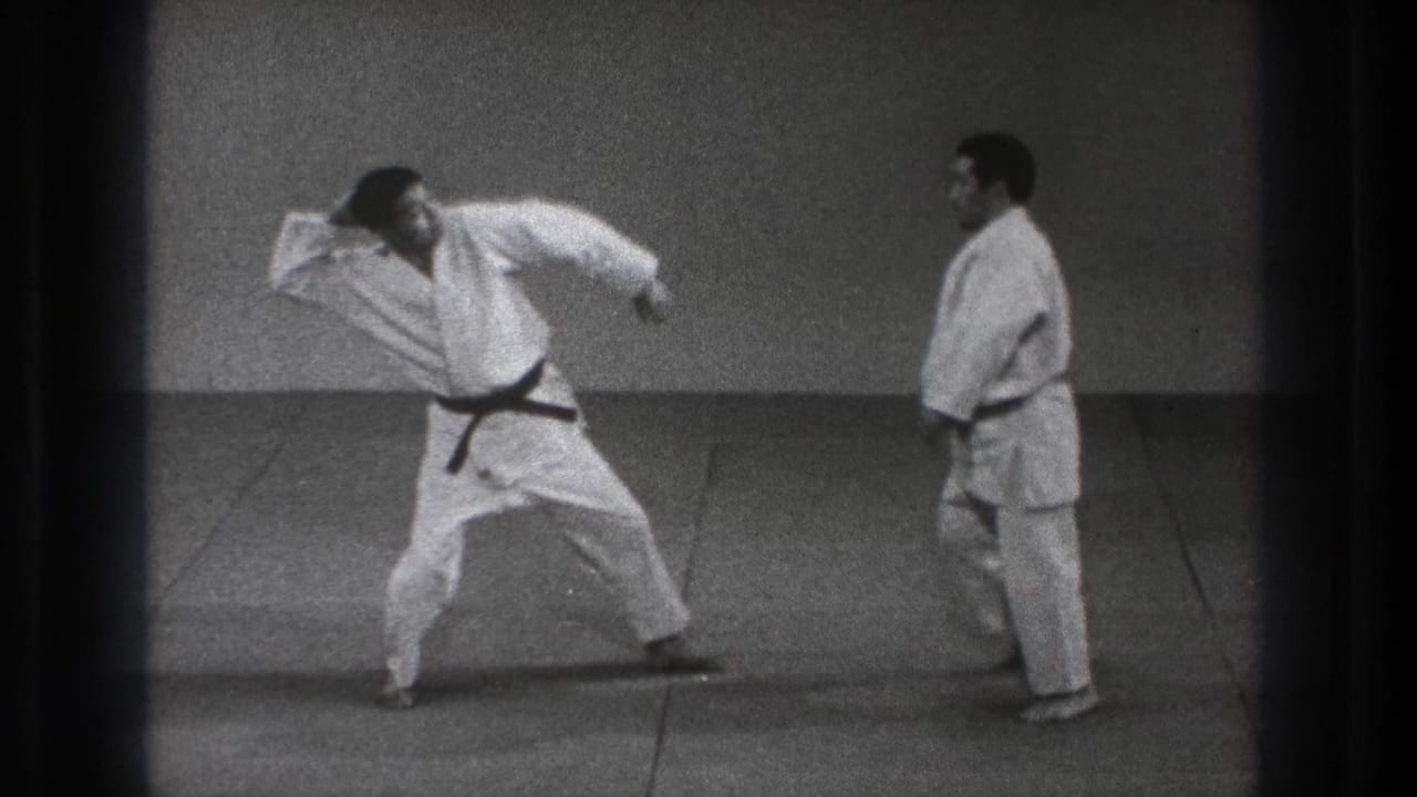 Two men in white gi stand on a mat as one holds a wide throwing stance.